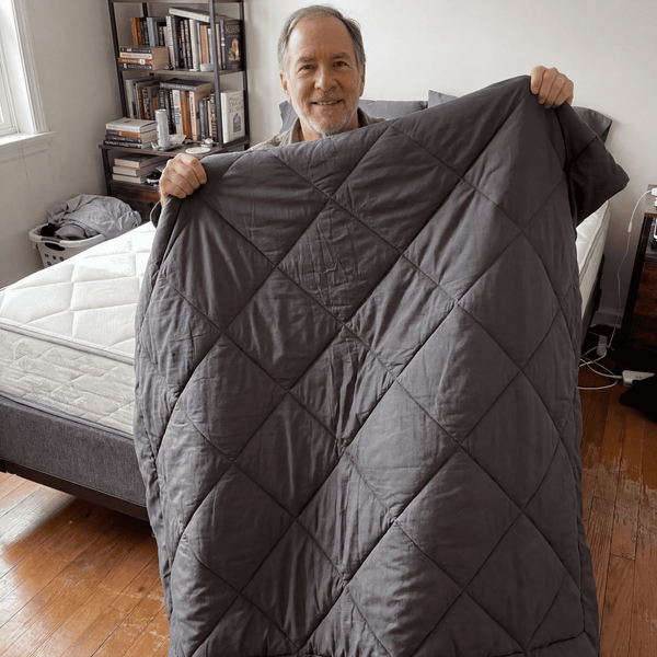 A customer holding up a gray 2-in-1 Comforter next to a bed